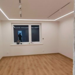 Bak-Pol Baka Krzysztof - Jasne wnętrze po remoncie: panele podłogowe, białe ściany, okno z widokiem na zewnątrz i nowoczesne oświetlenie LED na suficie. Częściowo wykończone ściany.