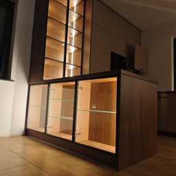 Bak-Pol Baka Krzysztof - Nowoczesna zabudowa meblowa z podświetlanymi szklanymi witrynami i drewnianymi frontami, widoczna na tle jasnej podłogi i ściany. Minimalistyczny design.