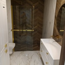 Bak-Pol Baka Krzysztof - Nowoczesna łazienka z kabiną prysznicową typu walk-in, białą szafką pod umywalką z złotymi uchwytami i marmurową podłogą. Ściany wykończone płytkami w odcieniach brązu i bieli.