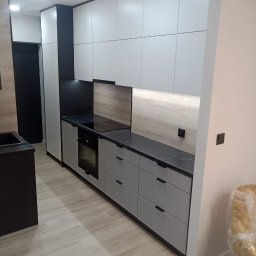 Bak-Pol Baka Krzysztof - Nowoczesna kuchnia z szarymi szafkami i czarnym blatem. Górne szafki białe, podświetlane. Widoczny fragment krzesła w folii. Minimalistyczny design.