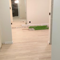 Bak-Pol Baka Krzysztof - Jasny parkiet w korytarzu, widoczne paczki paneli podłogowych Wenninger. W lustrze odbija się osoba. Ściany białe, minimalistyczne oświetlenie.