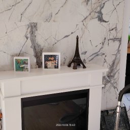 Bak-Pol Baka Krzysztof - Białe płytki imitujące marmur na ścianie za kominkiem elektrycznym. Dekoracje na kominku, widoczny fragment kuchni i krzesła. Użycie odkurzacza w trakcie sprzątania.