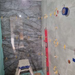 Bak-Pol Baka Krzysztof - Układanie płytek imitujących marmur na ścianie w łazience. Widoczny poziomica, drabina i system poziomowania płytek. Prace glazurnicze w toku.