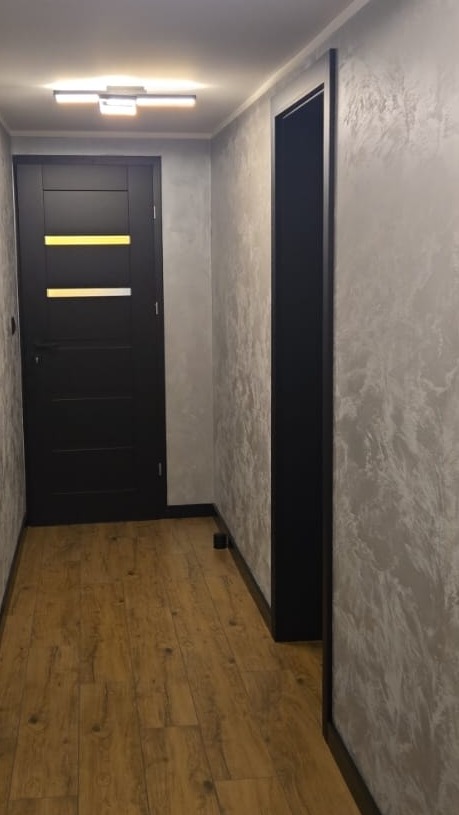 Korytarz po remoncie: drewnopodobna podłoga, ściany z efektem dekoracyjnym, czarne drzwi z poziomymi, żółtymi akcentami i czarna ościeżnica. Nowoczesne oświetlenie sufitowe.