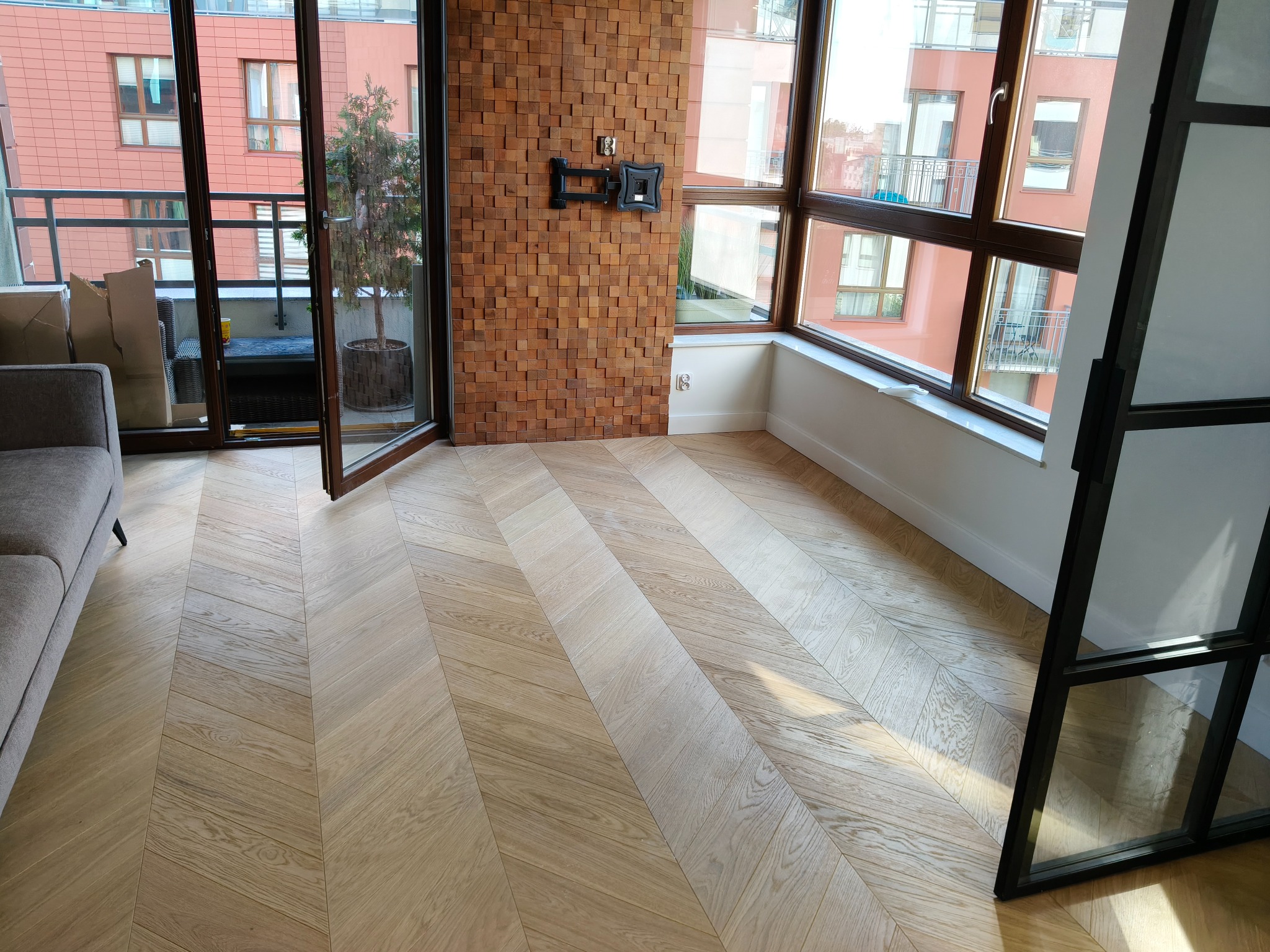 Jasny parkiet ułożony we wzór jodełki francuskiej w nowoczesnym salonie z wyjściem na balkon. Na ścianie drewniana mozaika i uchwyt na telewizor.