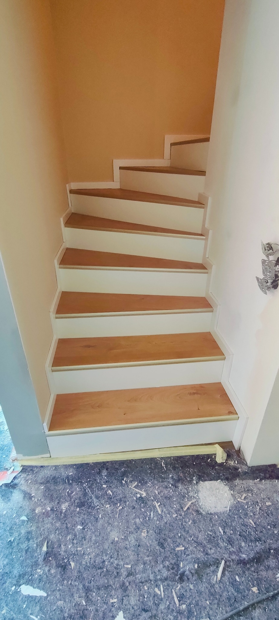 Wykończone schody z drewnianymi stopniami i białymi podstopnicami. Widoczne detale wykończenia i zabezpieczona podłoga. Ujęcie z dołu schodów.