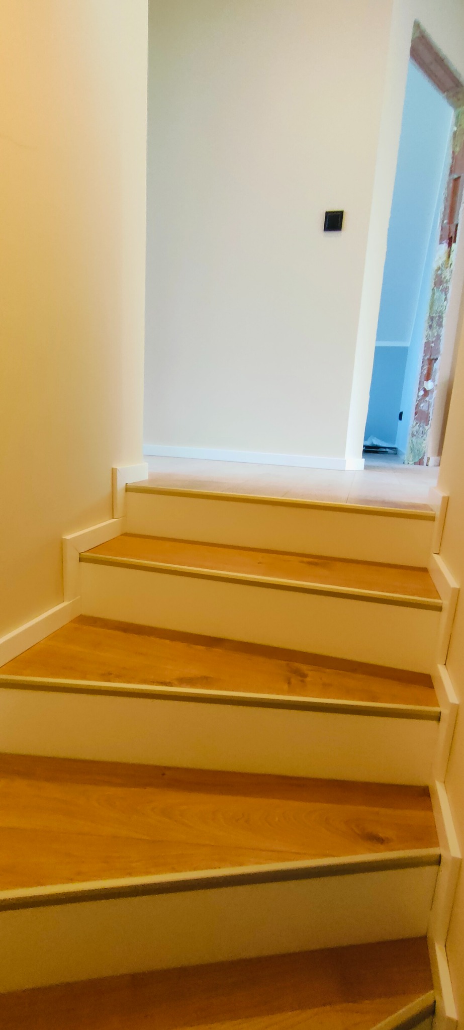 Wykończone schody z drewnianymi stopniami i białymi podstopnicami, zabezpieczone metalowymi listwami. Widok z dołu, jasne ściany i fragment korytarza w tle.