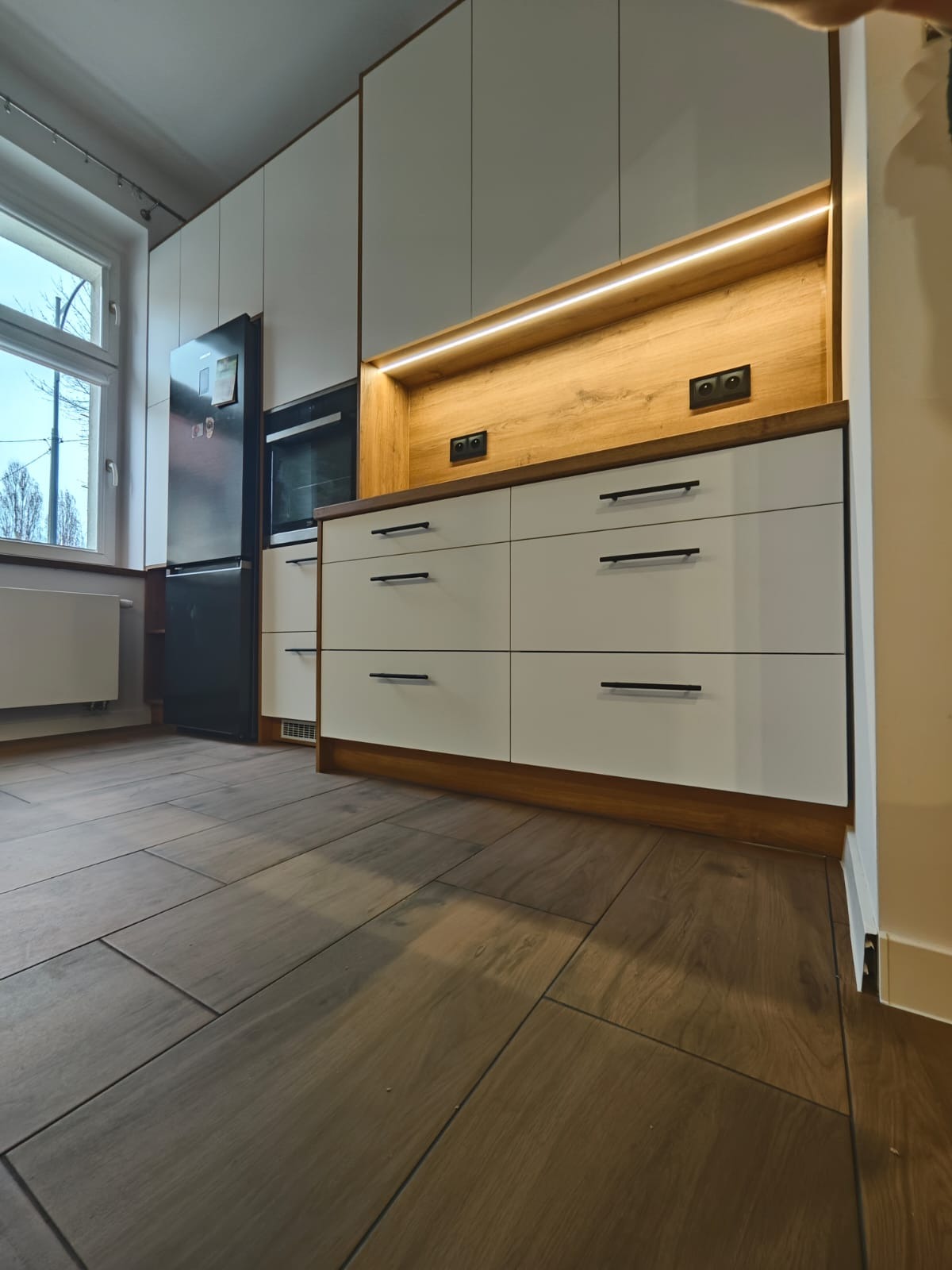 Nowoczesna zabudowa kuchenna z oświetleniem LED w Tczewie. Białe fronty szafek i drewnopodobne wykończenia. Widok na okno i podłogę z płytek imitujących drewno.