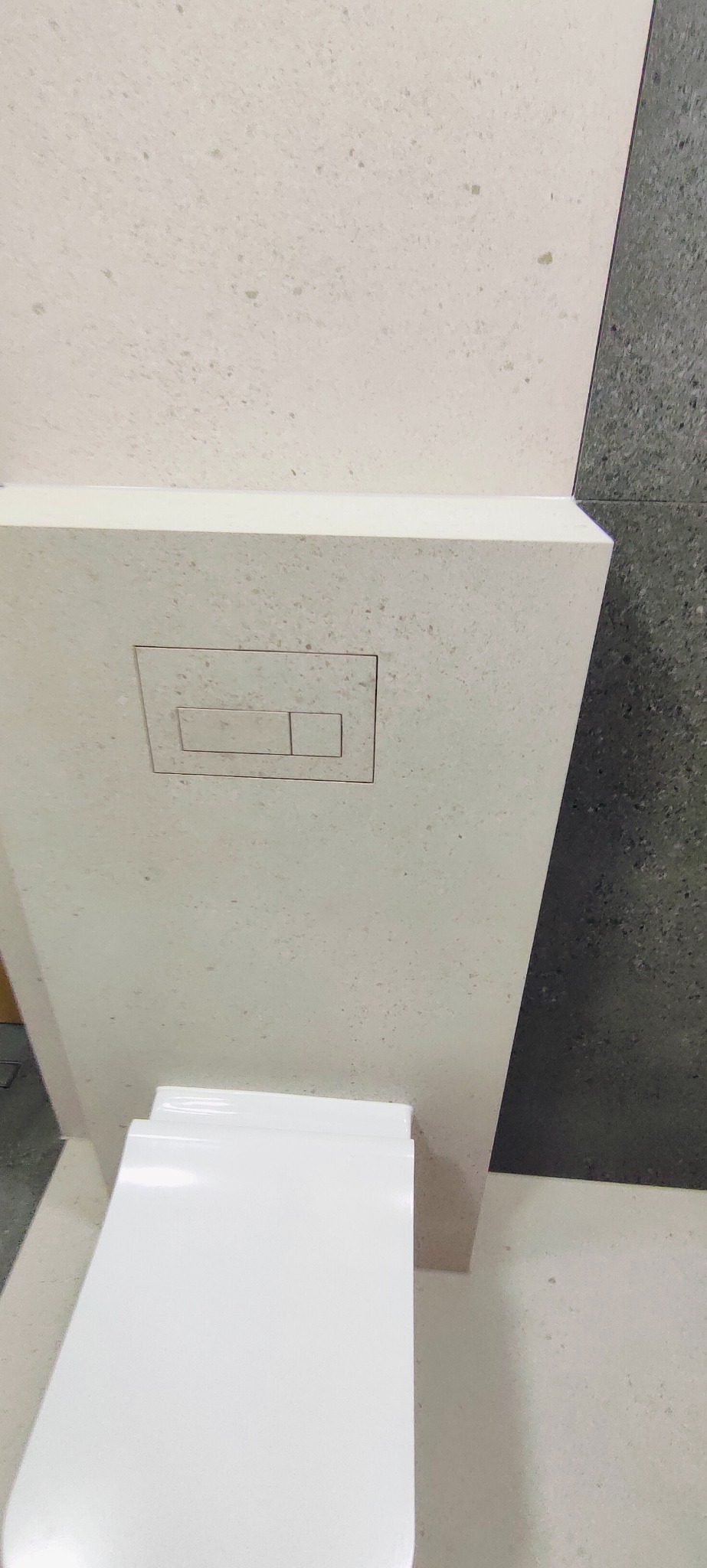Kompakt WC z zabudową z jasnego kamienia z przyciskiem spłukującym, na tle ściany z ciemnych płytek. Minimalistyczny design, nowoczesna łazienka.