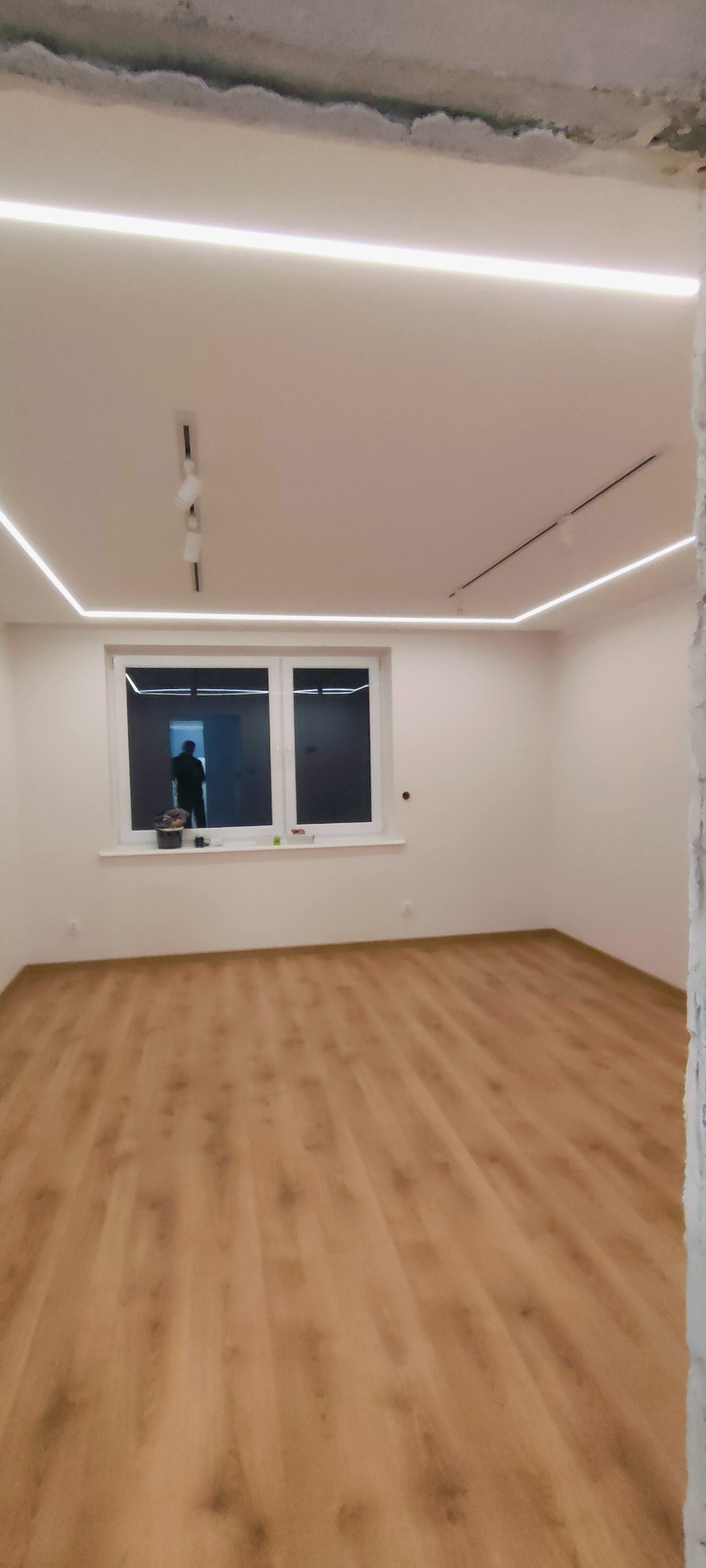 Jasne wnętrze po remoncie: panele podłogowe, białe ściany, okno z widokiem na zewnątrz i nowoczesne oświetlenie LED na suficie. Częściowo wykończone ściany.