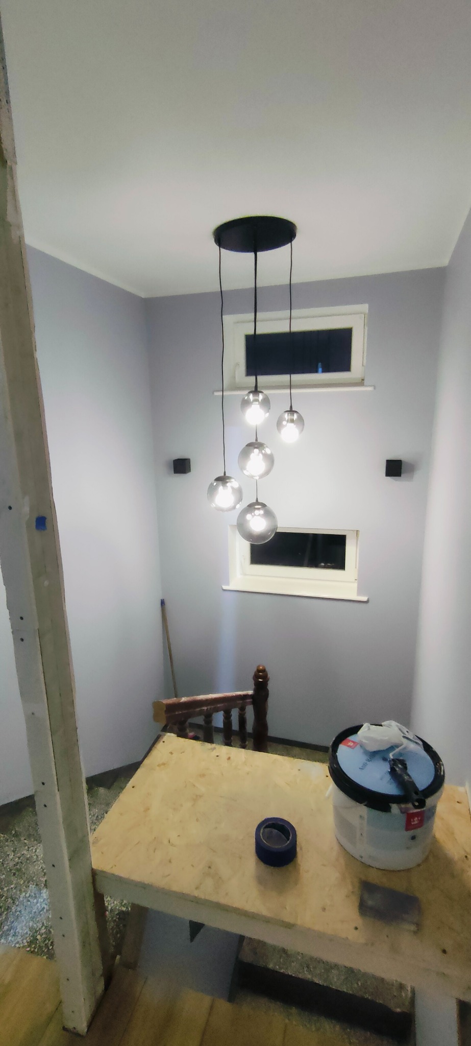 Jasne wnętrze w trakcie malowania: ściany w odcieniach fioletu, nowoczesna lampa wisząca z kulistymi kloszami, okna i narzędzia malarskie na prowizorycznym stole.