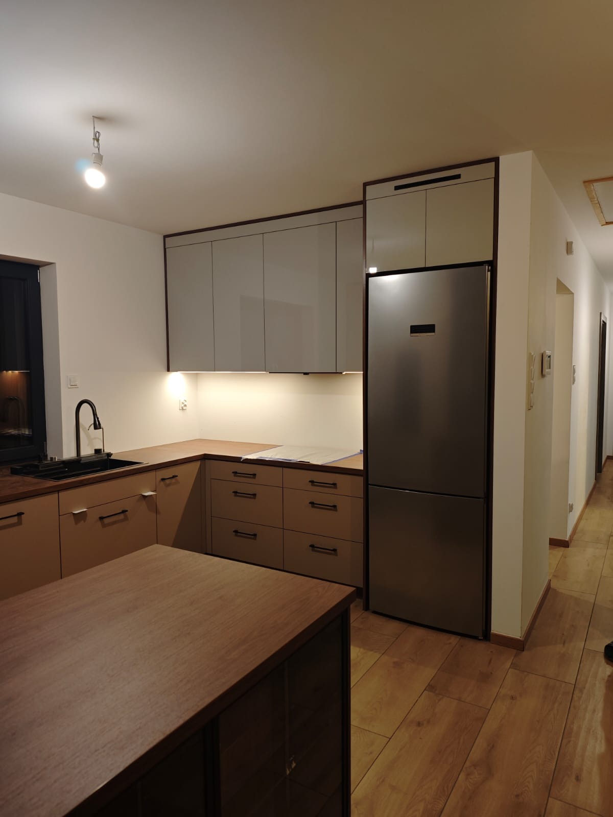 Nowoczesna kuchnia z wyspą: beżowe szafki dolne, szare górne, stalowa lodówka. Ustawienie w kształcie litery L, oświetlenie podszafkowe, drewniana podłoga.