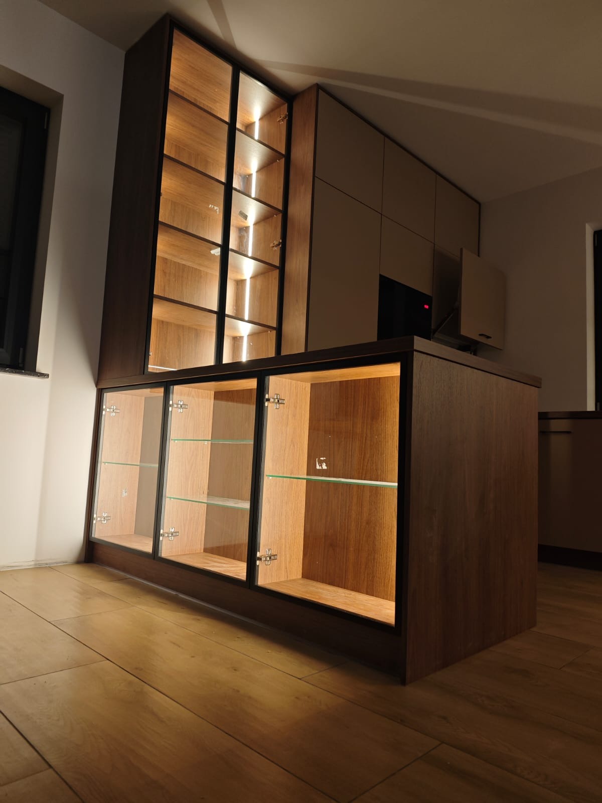 Nowoczesna zabudowa meblowa z podświetlanymi szklanymi witrynami i drewnianymi frontami, widoczna na tle jasnej podłogi i ściany. Minimalistyczny design.