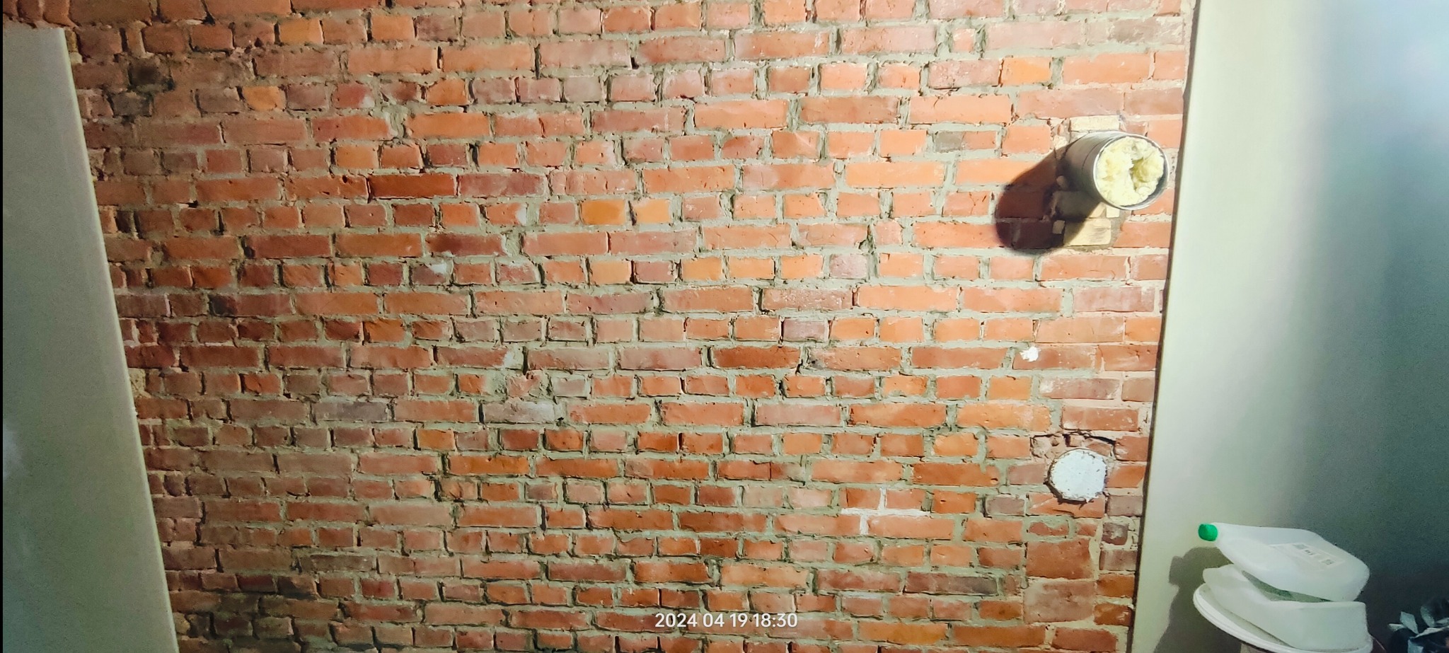 Ceglana ściana z widocznymi otworami instalacyjnymi i fragmentem ściany pomalowanej na jasny kolor. Widoczne ślady po instalacjach.