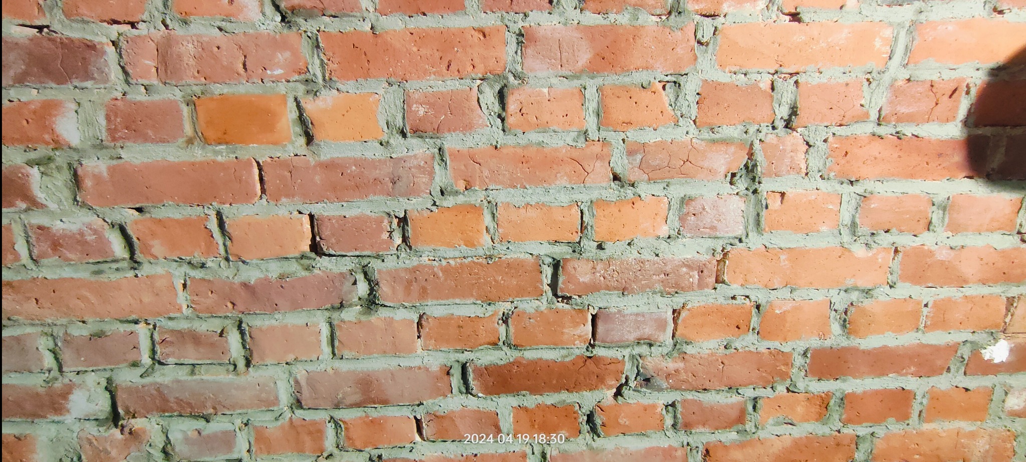Czerwona ściana z cegły z widoczną zaprawą, oświetlona z góry, z delikatnym cieniem w prawym górnym rogu. Widoczne nierówności i pęknięcia na cegłach.