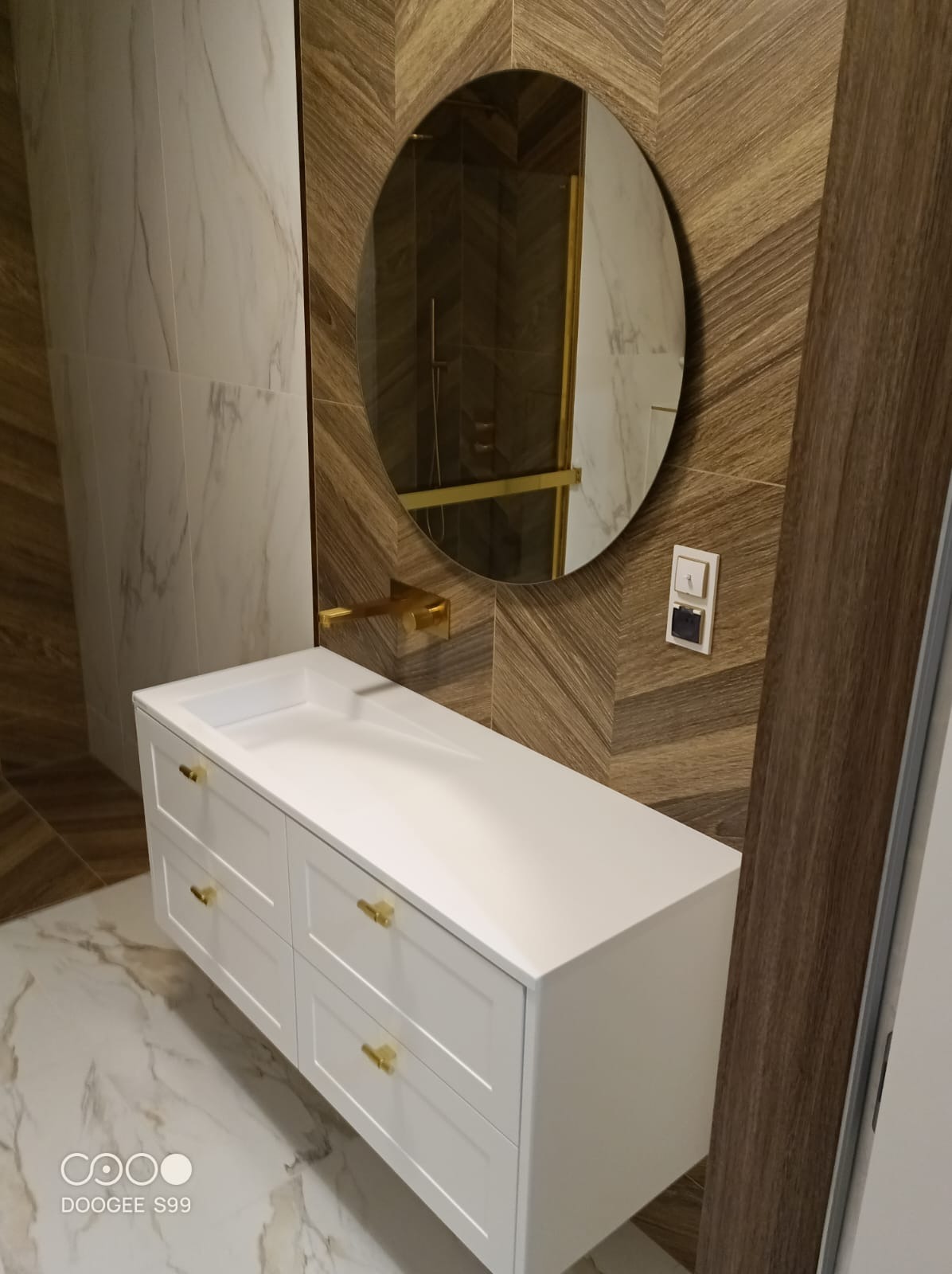 Nowoczesna łazienka: biała szafka z umywalką i złotymi uchwytami, owalne lustro z podświetleniem, wzorzyste płytki na ścianie i marmurowa podłoga.