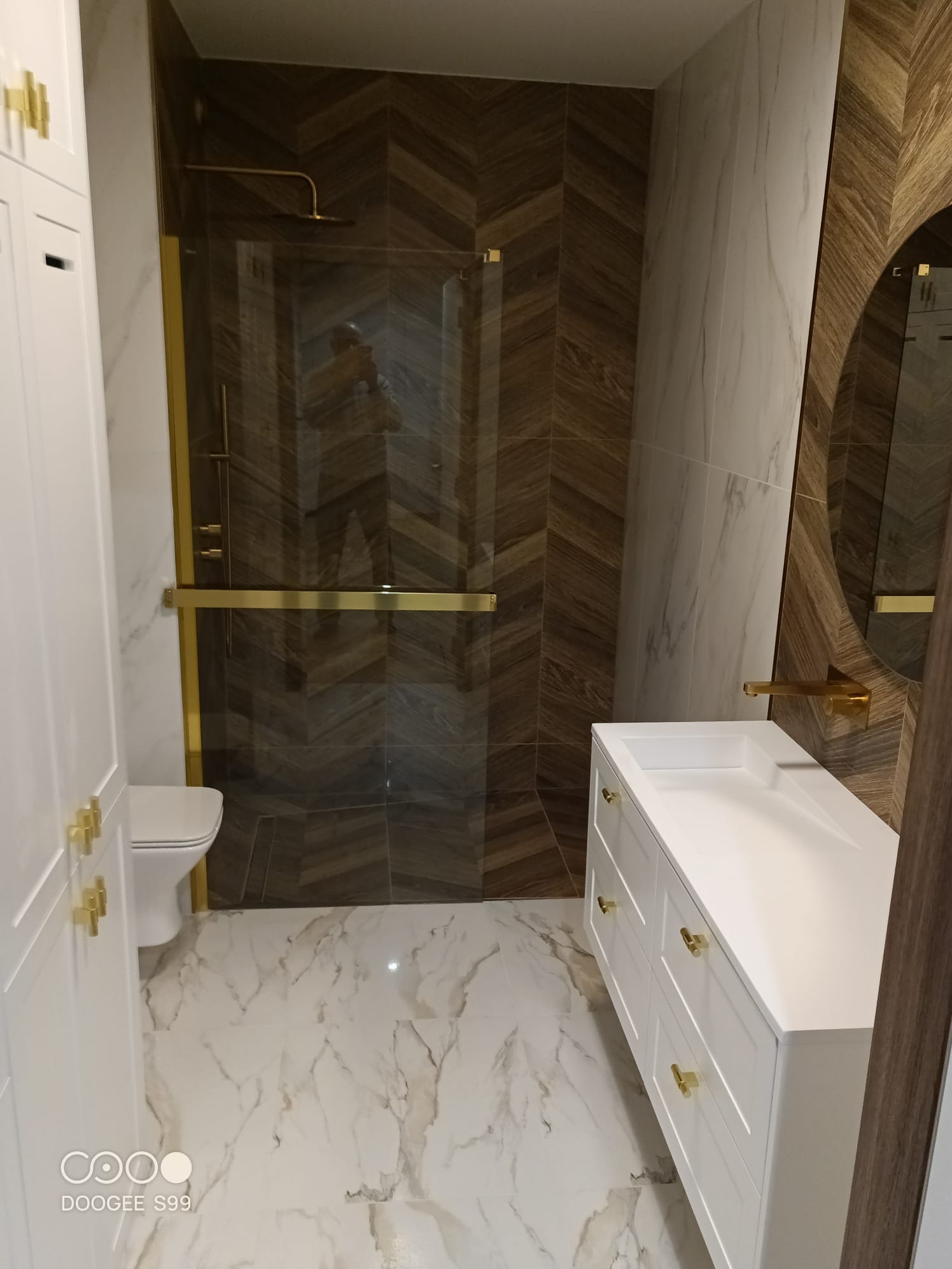 Nowoczesna łazienka z kabiną prysznicową typu walk-in, białą szafką pod umywalką z złotymi uchwytami i marmurową podłogą. Ściany wykończone płytkami w odcieniach brązu i bieli.