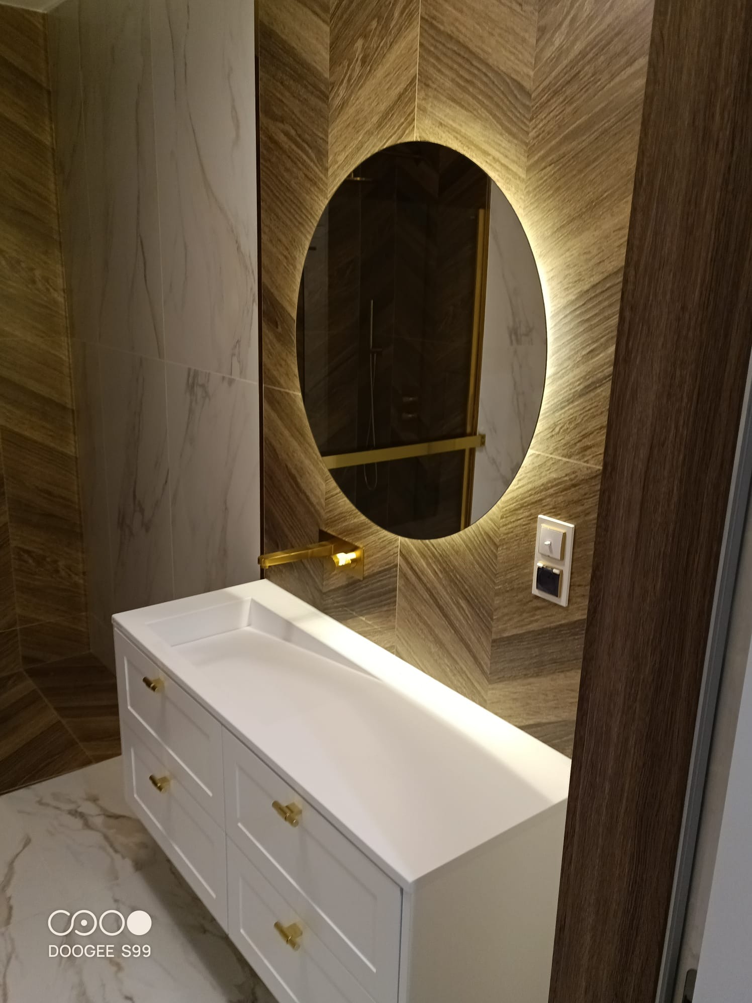 Nowoczesna łazienka z owalnym lustrem podświetlanym LED, białą szafką z umywalką i złotymi uchwytami. Na ścianie płytki ułożone w jodełkę i marmurowe.