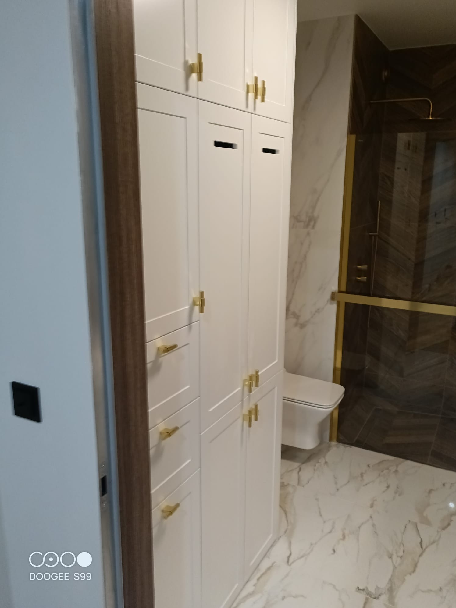 Biała zabudowa meblowa z złotymi uchwytami w łazience z marmurową podłogą i kabiną prysznicową w tle. Widok na toaletę i fragment ściany.