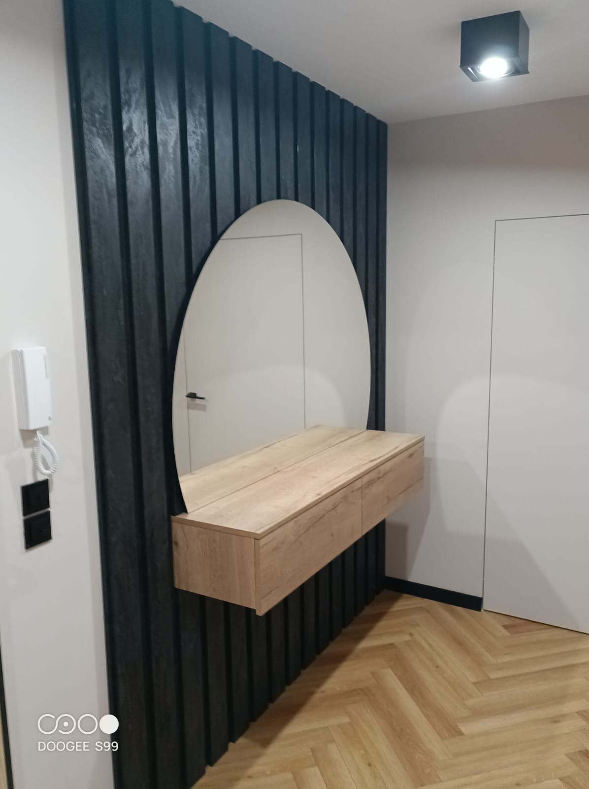 Nowoczesna aranżacja przedpokoju: owalne lustro z drewnianą konsolą na tle ściany z ciemnych, pionowych paneli. Parkiet w jodełkę, minimalistyczne drzwi i oświetlenie.