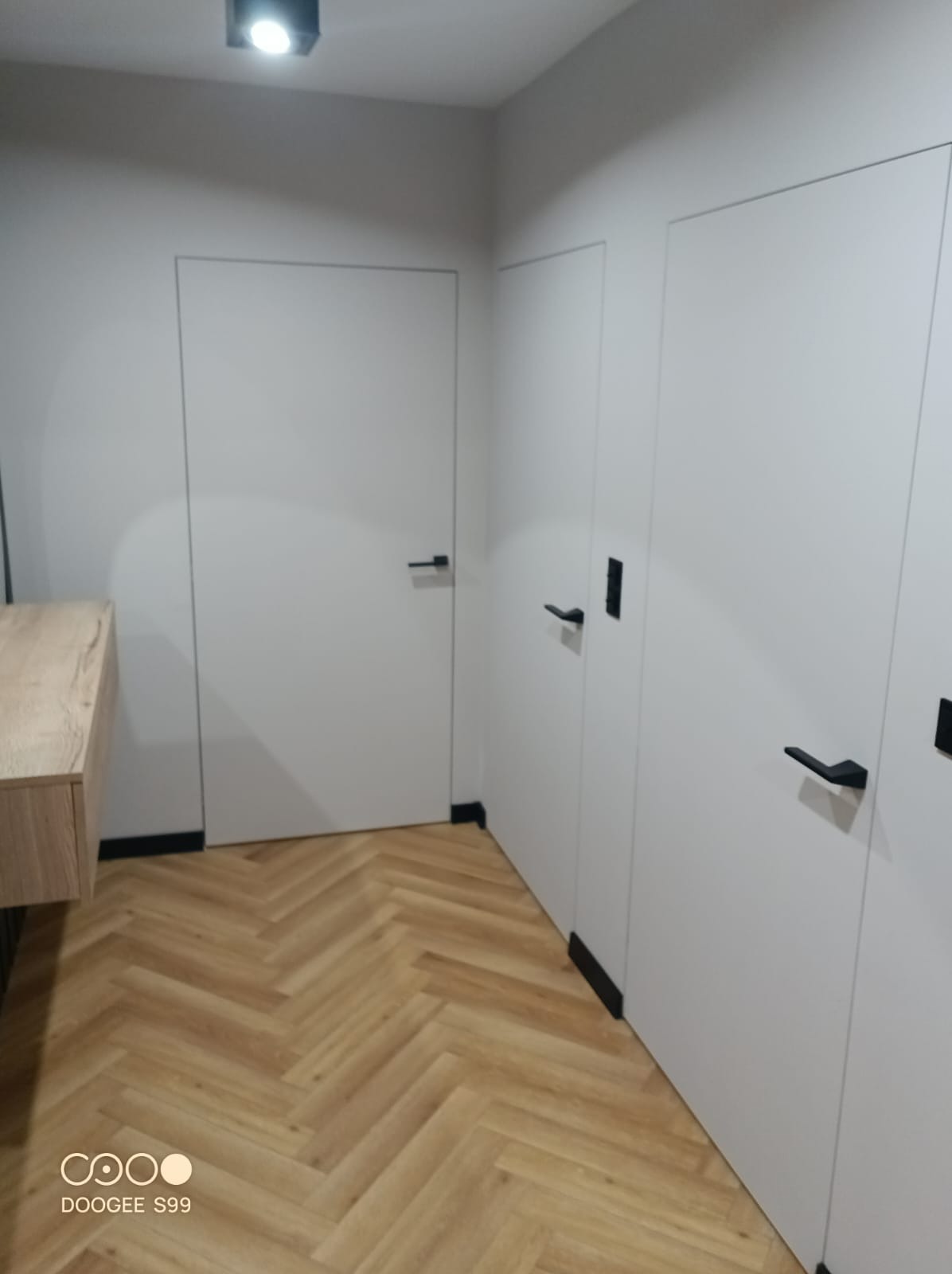 Nowoczesne wnętrze: parkiet w jodełkę, białe drzwi z czarnymi klamkami, minimalistyczna szafka. Oświetlenie sufitowe. Widok na wykończony korytarz.