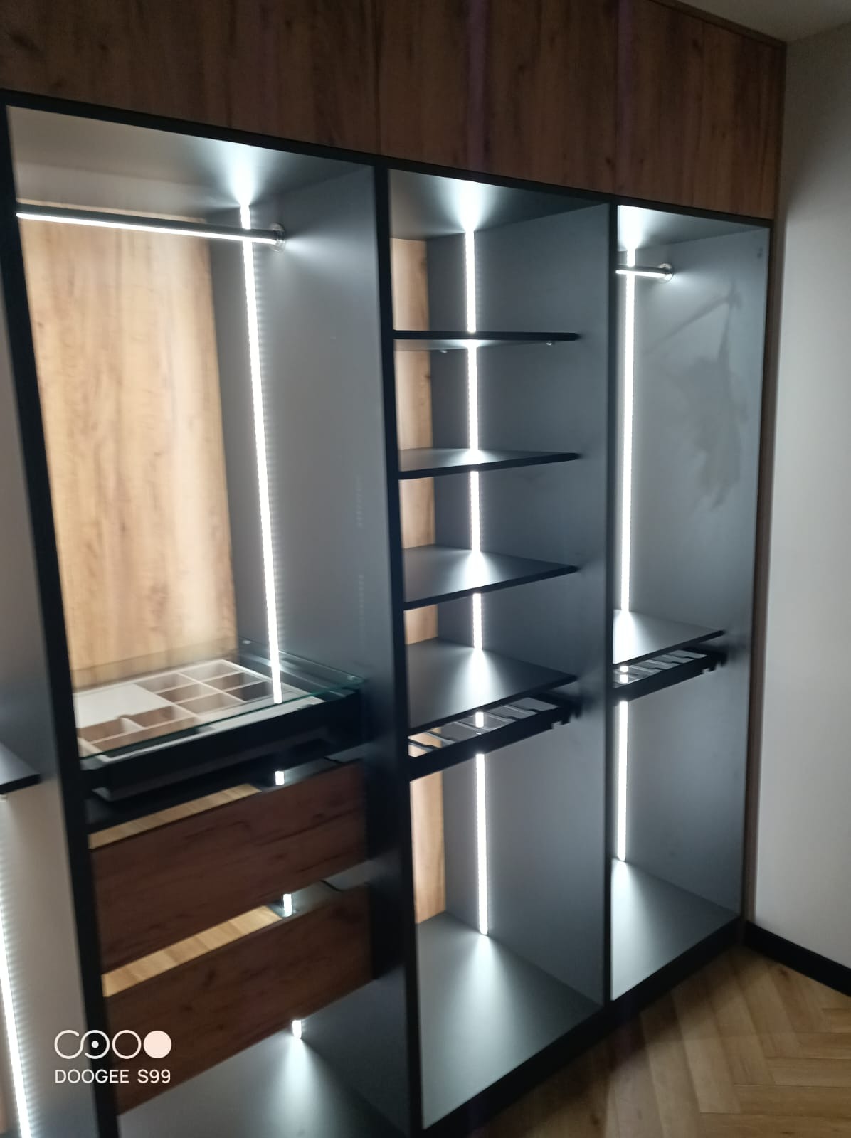 Nowoczesna, otwarta garderoba z szarymi półkami, drewnianymi elementami i oświetleniem LED. Widoczne szuflady i półki na akcesoria. Podłoga parkiet.