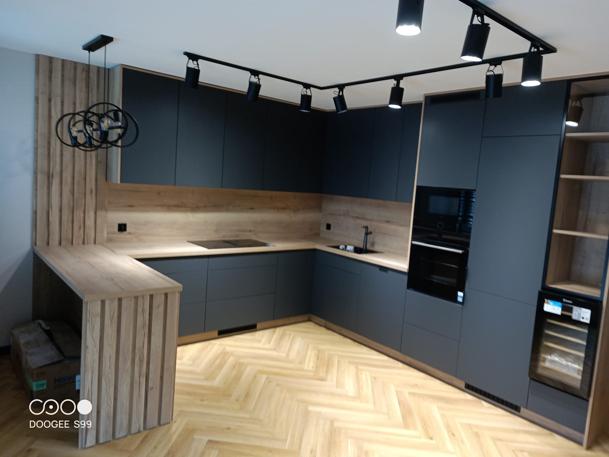 Nowoczesna kuchnia w odcieniach szarości z drewnianymi akcentami, oświetleniem punktowym i parkietem ułożonym w jodełkę. Widoczny barek i sprzęt AGD.