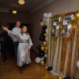 Uczestnicy zabawy na tle dekoracji z balonów i cyfr 55 w kolorach złota, bieli i czerni. Kobieta w srebrnej sukience tańczy na parkiecie. Uroczystość w sali.