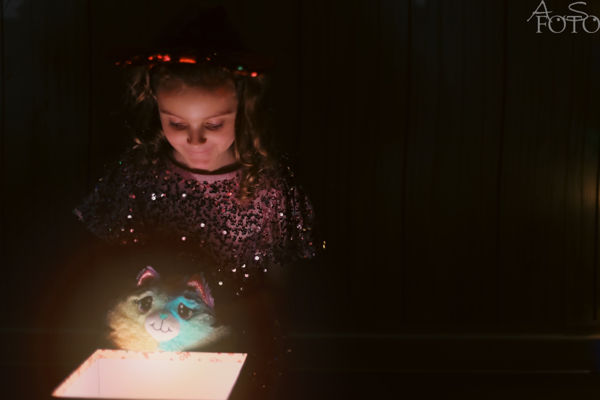 Dziewczynka w cekinowej bluzce z zaciekawieniem spogląda w dół na otwarte pudełko, z którego wydobywa się jasne światło, oświetlając jej twarz i pluszową zabawkę.