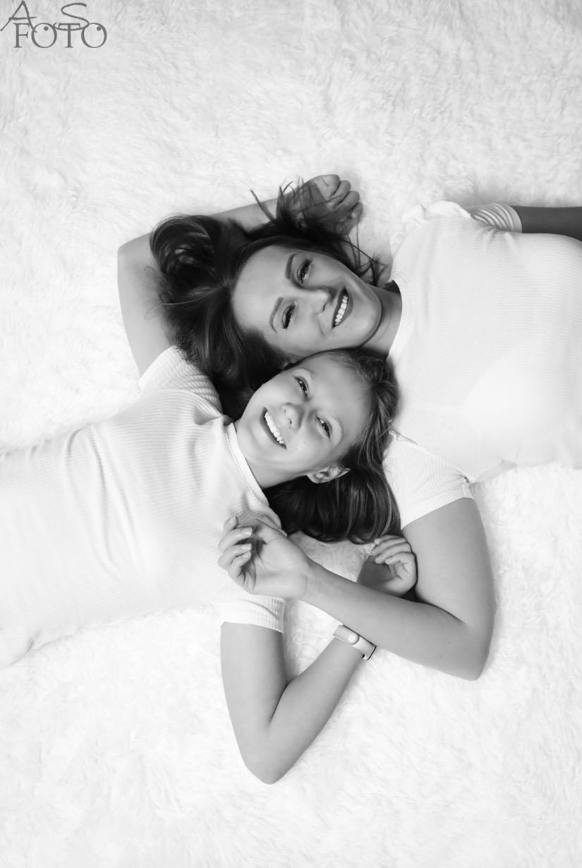 Czarno-białe zdjęcie matki i córki leżących na białym, puszystym dywanie, uśmiechniętych, z widocznym logo fotografa w lewym górnym rogu. Ujęcie z góry.