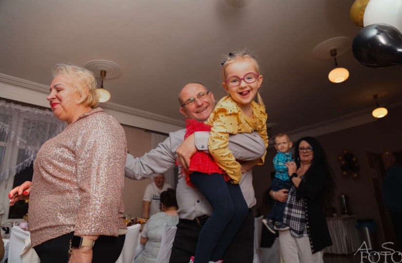 Uśmiechnięta dziewczynka w okularach na rękach mężczyzny, w tle baloniki i goście na przyjęciu. Fotografia z uroczystości rodzinnej.