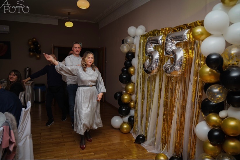 Uczestnicy zabawy na tle dekoracji z balonów i cyfr 55 w kolorach złota, bieli i czerni. Kobieta w srebrnej sukience tańczy na parkiecie. Uroczystość w sali.