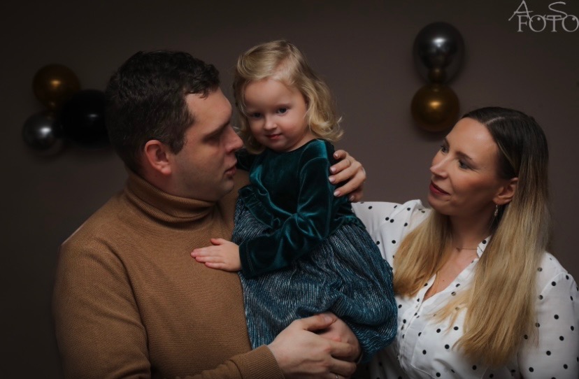 Uśmiechnięta rodzina: tata trzyma córeczkę w zielonej sukience, mama patrzy z boku. W tle balony i logo fotografa. Ciepłe, naturalne światło.