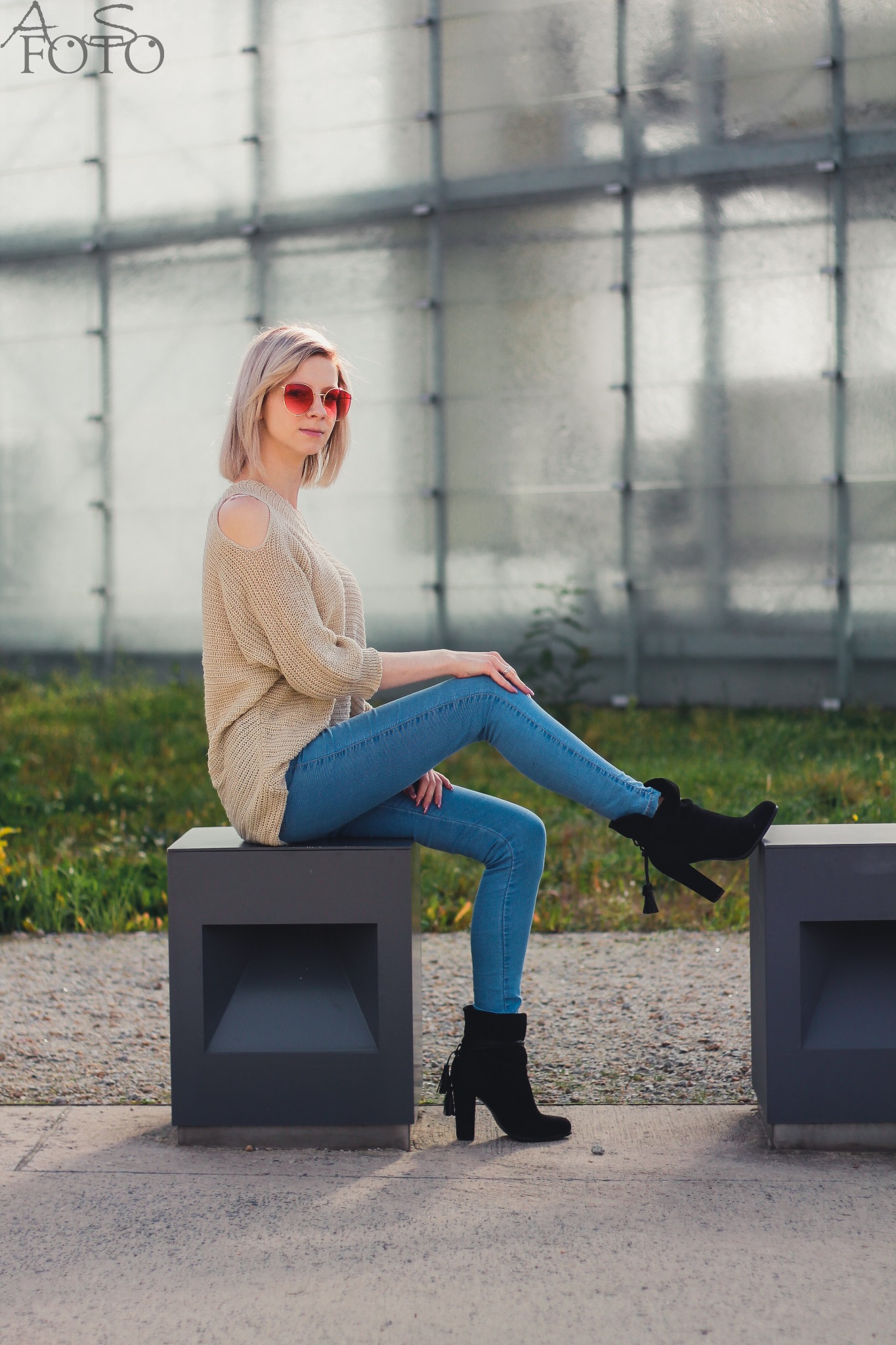 Blondynka w swetrze z odkrytym ramieniem i czerwonych okularach siedzi na betonowym sześcianie na tle szklanej ściany. Ubrana w dżinsy i czarne botki na obcasie.
