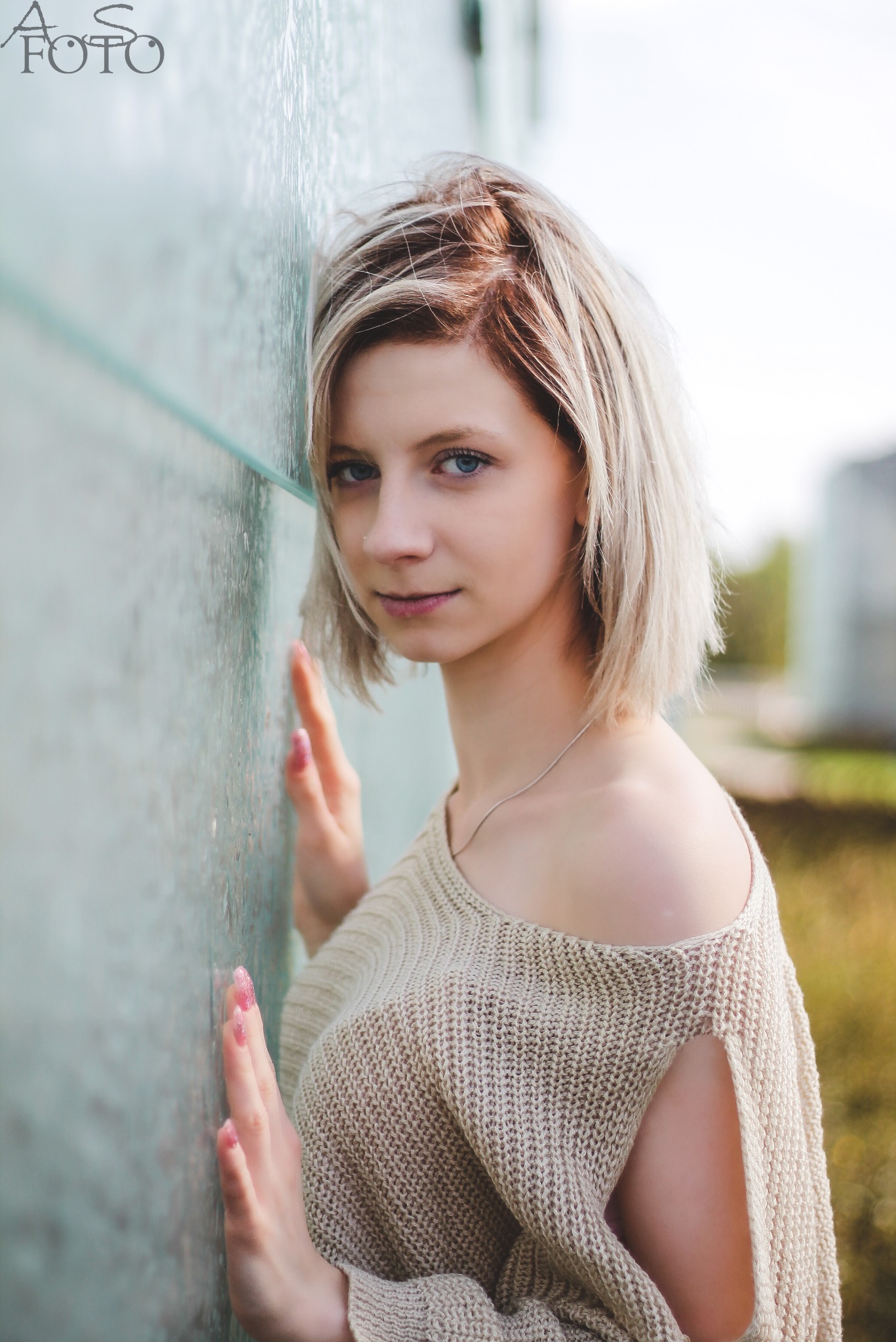 Portret młodej kobiety z blond włosami, opierającej się o szklaną ścianę. Ubrana w beżowy sweter z odkrytym ramieniem, delikatny uśmiech, naturalne światło.