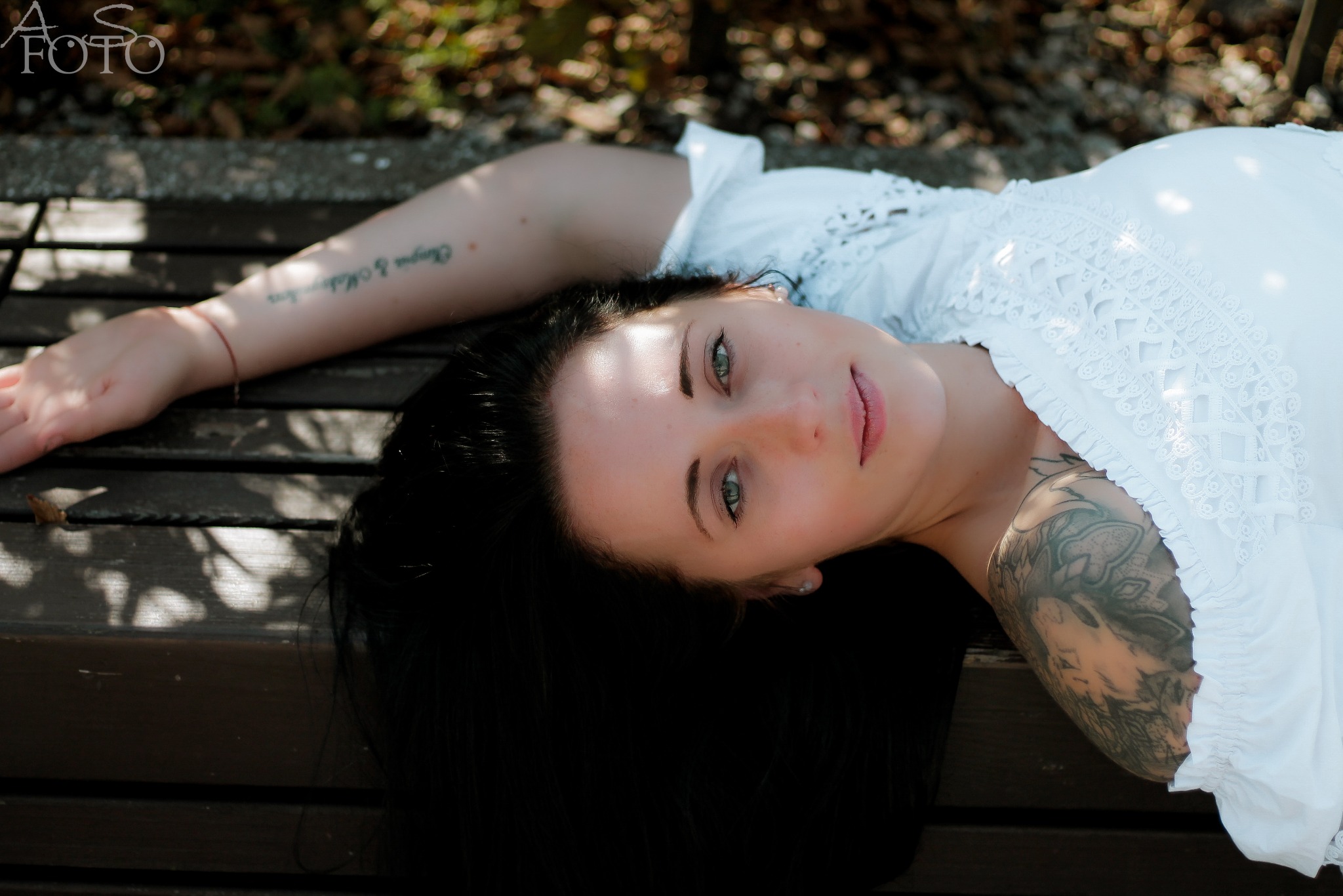Kobieta w białej sukience leży na ławce, z tatuażami na ramionach, patrzy w obiektyw. Ujęcie z góry, naturalne światło podkreśla zielone oczy.