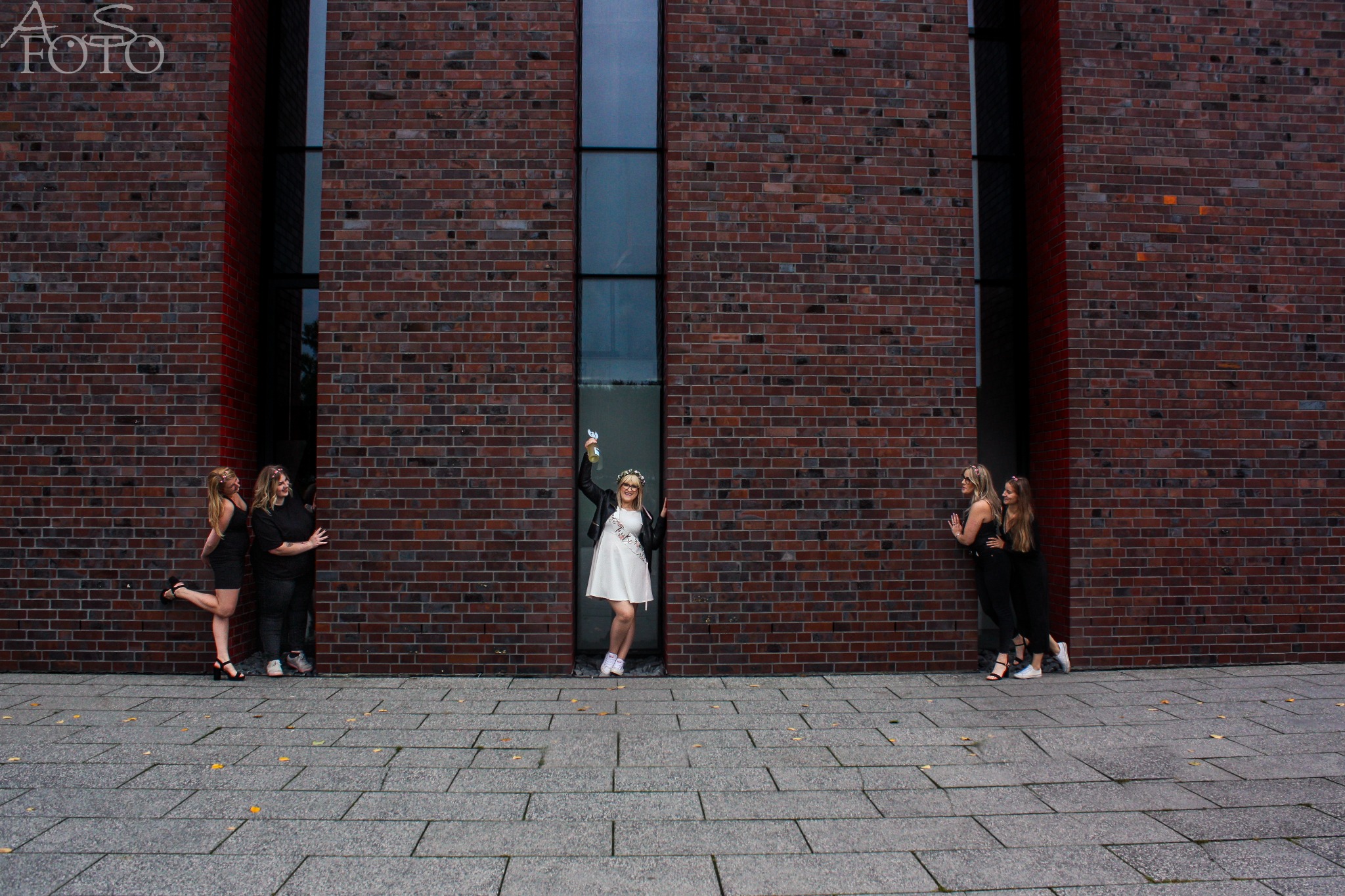 Panna młoda w białej sukience i wianku, z butelką w ręku, pozuje z przyjaciółkami na tle ceglanej ściany z wąskimi oknami. Ujęcie z dołu, szeroki kadr.