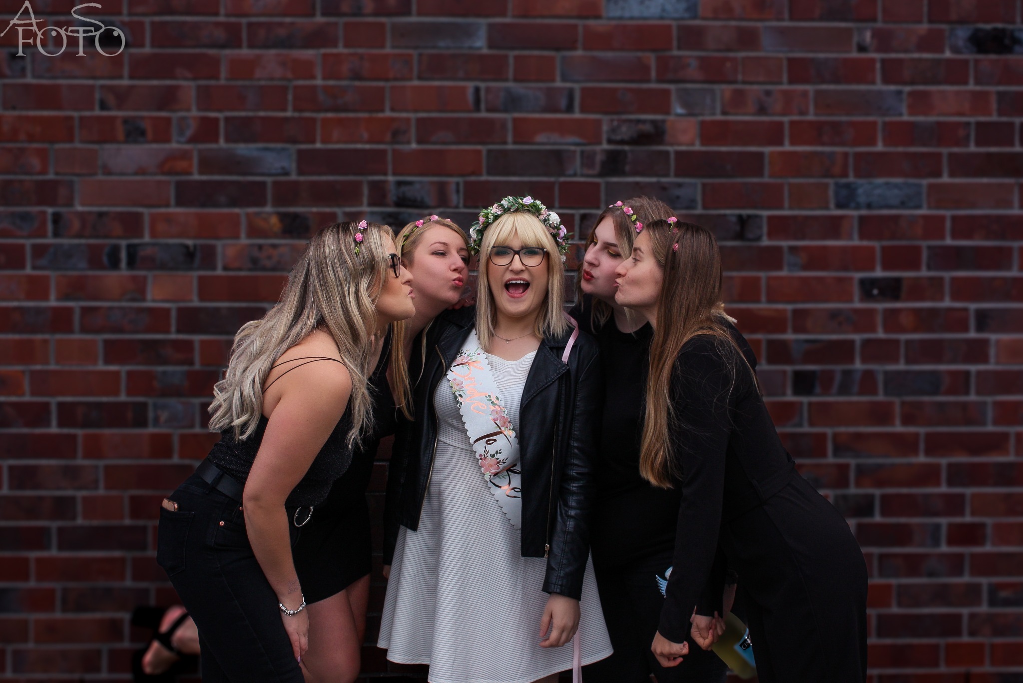 Uśmiechnięta blondynka w okularach i wianku otoczona przez cztery całujące ją przyjaciółki na tle ceglanej ściany. Wieczór panieński.