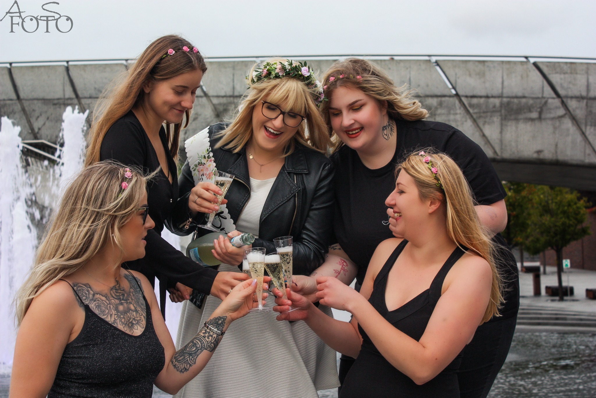 Grupa pięciu uśmiechniętych kobiet w kwiecistych wiankach wznosi toast szampanem na tle fontanny i betonowego mostu. Jedna z kobiet ma na sobie skórzaną kurtkę.
