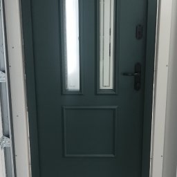 FUZION MPL SPÓŁKA Z OGRANICZONĄ ODPOWIEDZIALNOŚCIĄ - Ciemnozielone drzwi garażowe z dwoma pionowymi okienkami i czarną klamką, zamontowane w białej ramie. Widoczny fragment prowadnicy garażowej.