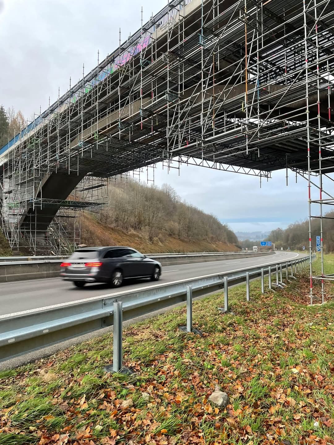 Most drogowy w trakcie remontu, otoczony rusztowaniami. Na pierwszym planie przejeżdżający samochód na autostradzie, jesienne liście na poboczu. Pochmurne niebo w tle.