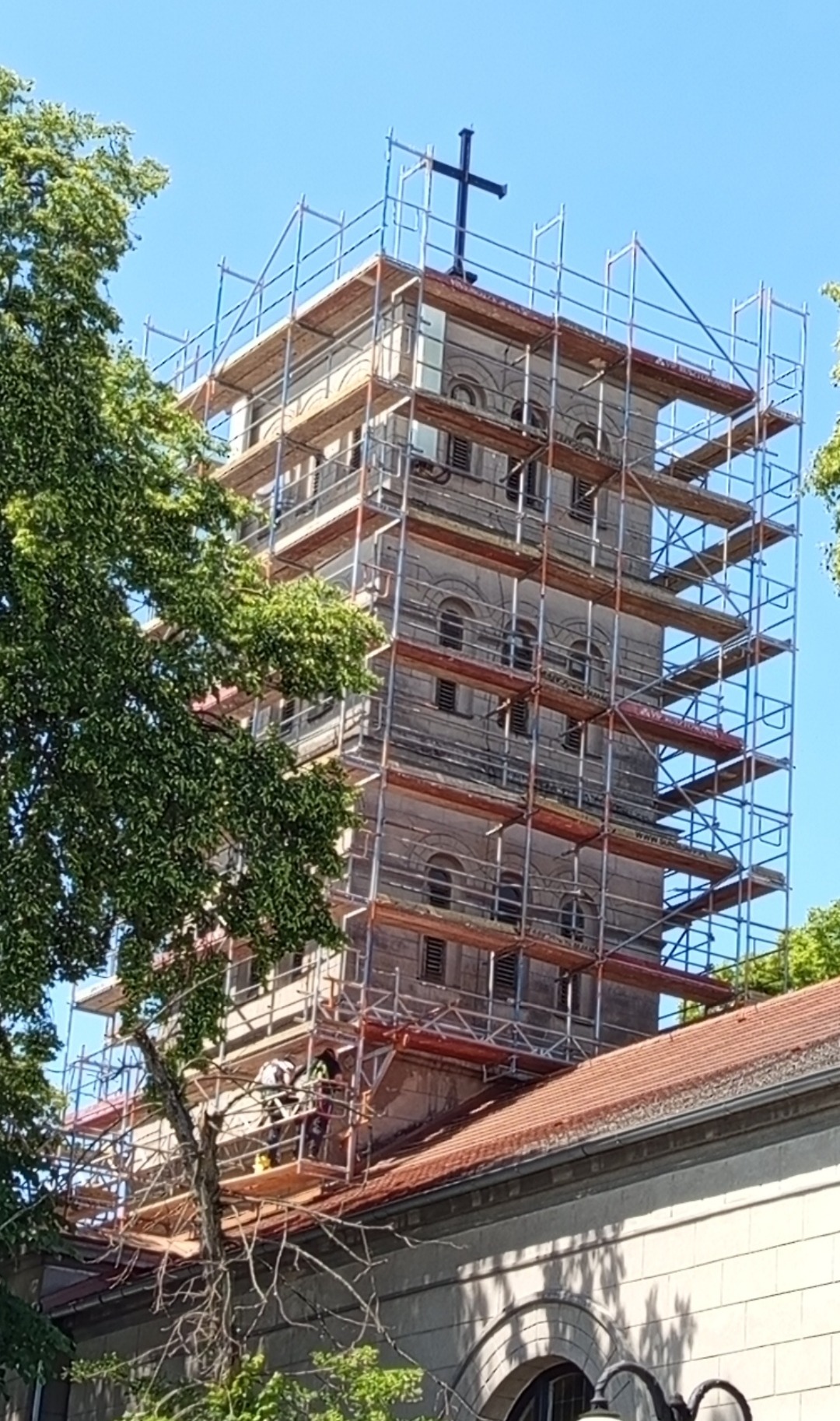 Wieża kościoła w rusztowaniach, prace renowacyjne w toku. Widoczni pracownicy na rusztowaniu. Błękitne niebo w tle, częściowo zasłonięte przez drzewa.