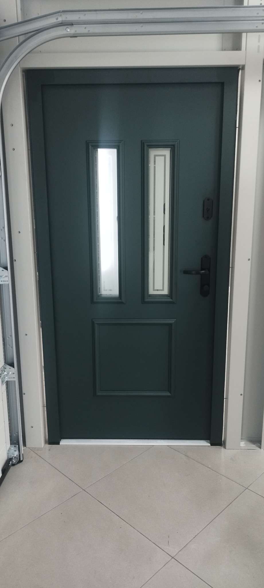 Ciemnozielone drzwi garażowe z dwoma pionowymi okienkami i czarną klamką, zamontowane w białej ramie. Widoczny fragment prowadnicy garażowej.