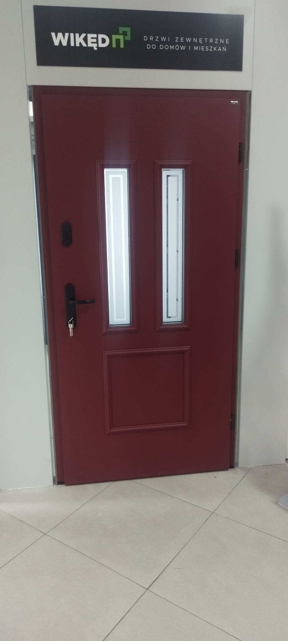 Bordowe drzwi zewnętrzne z szybkami i czarną klamką, klucz w zamku, pod szyldem 'WIKĘD - drzwi zewnętrzne do domów i mieszkań'.