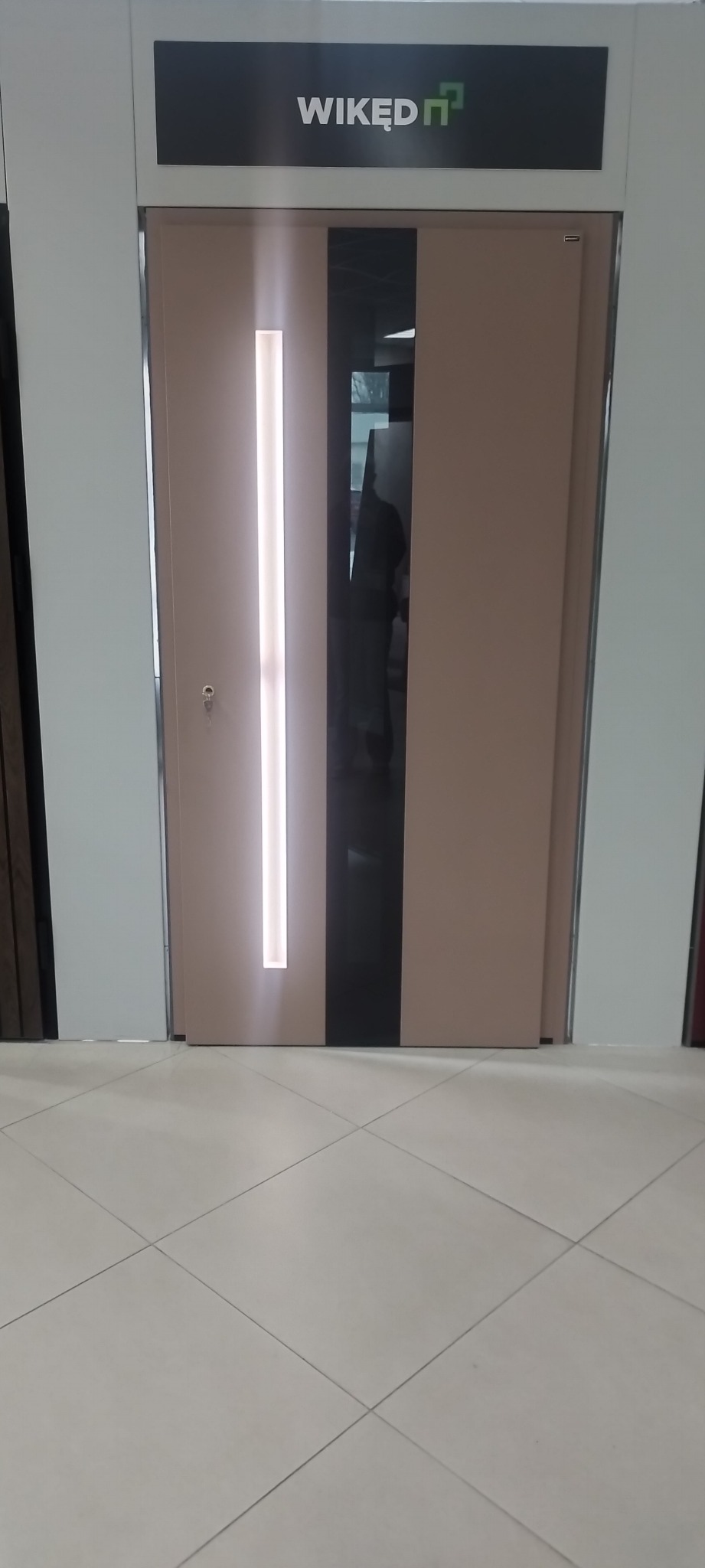 Beżowe drzwi wejściowe z podłużnym oświetleniem LED i czarnym panelem, widoczne logo producenta WIKĘD na górze. Prezentacja w salonie.