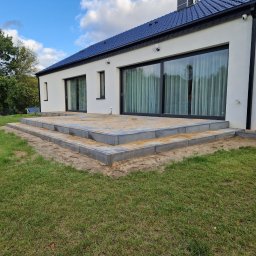 Martyna Sobczyńska - Nowoczesny dom z tarasem w trakcie budowy. Widoczne szare płyty tarasowe i świeżo ułożona kostka brukowa. Biała elewacja budynku z dużymi oknami przesuwnymi.