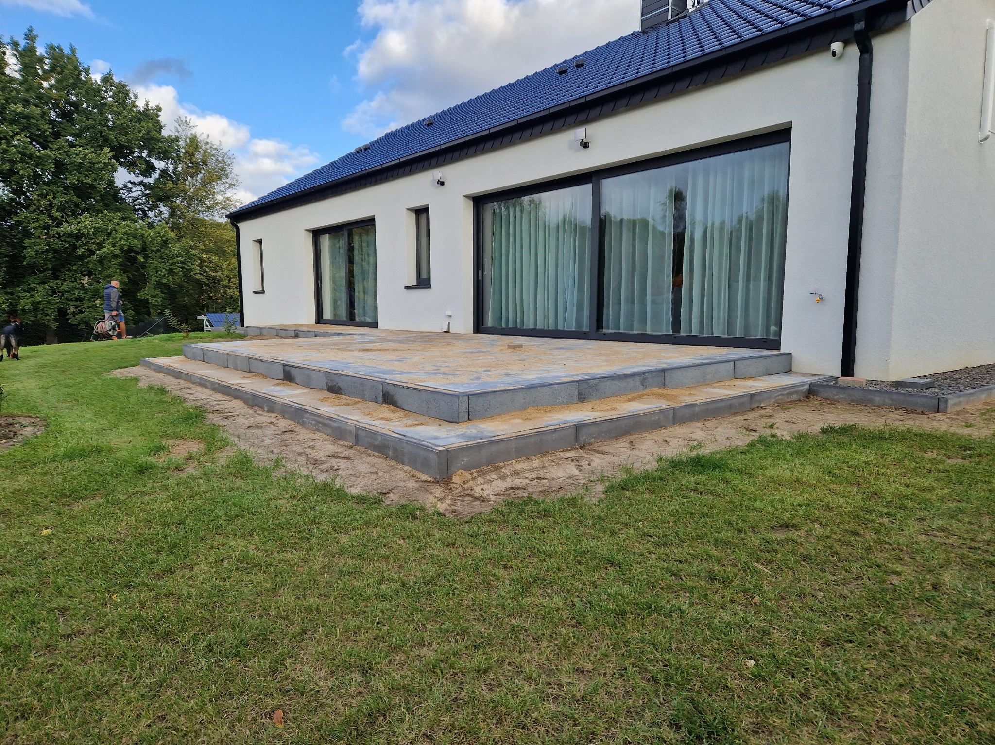 Nowoczesny dom z tarasem w trakcie budowy. Widoczne szare płyty tarasowe i świeżo ułożona kostka brukowa. Biała elewacja budynku z dużymi oknami przesuwnymi.