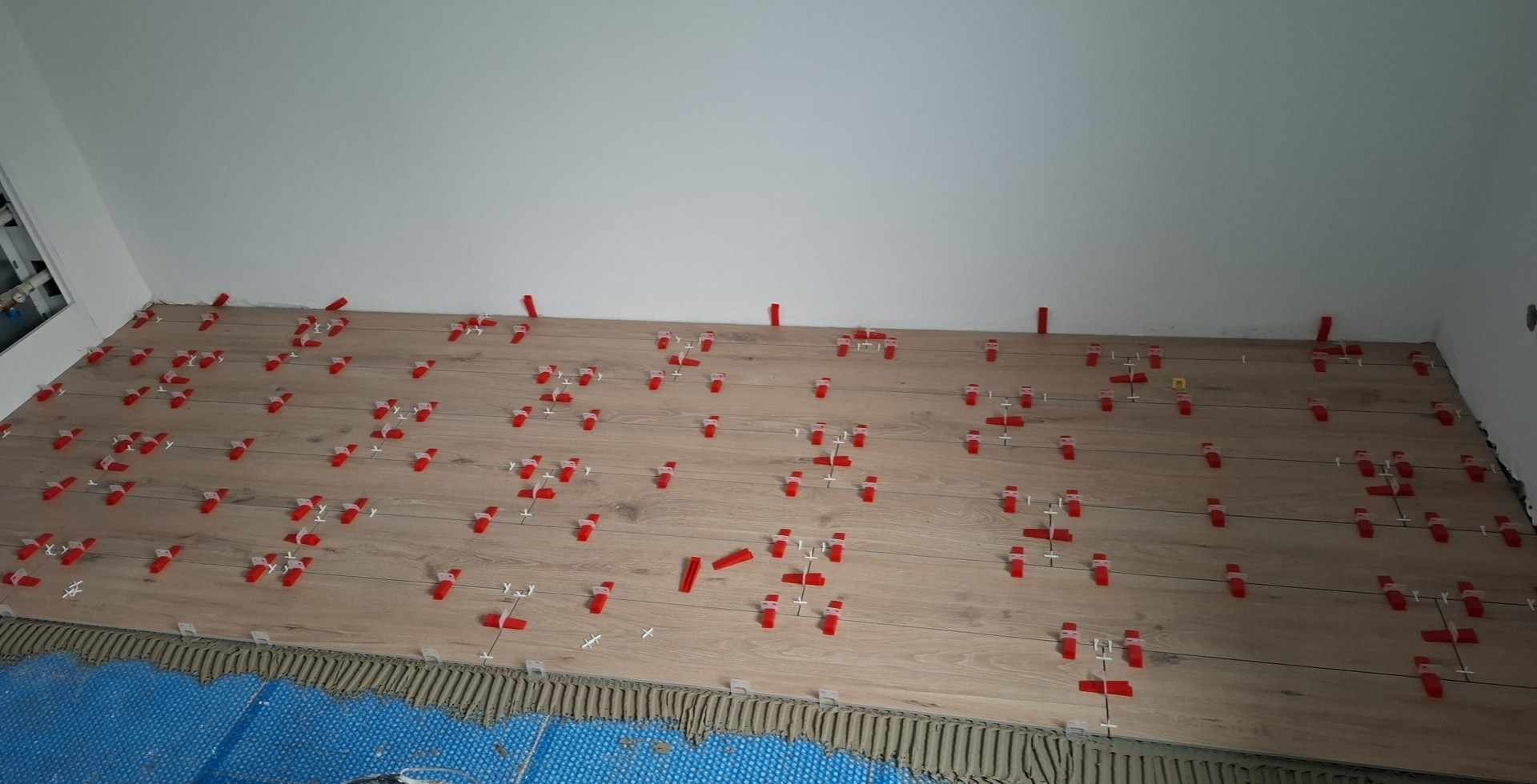 Układanie płytek imitujących drewno na podłodze. Widoczne czerwone klipsy poziomujące i niebieska mata podkładowa. Świeżo położona podłoga w trakcie prac wykończeniowych.