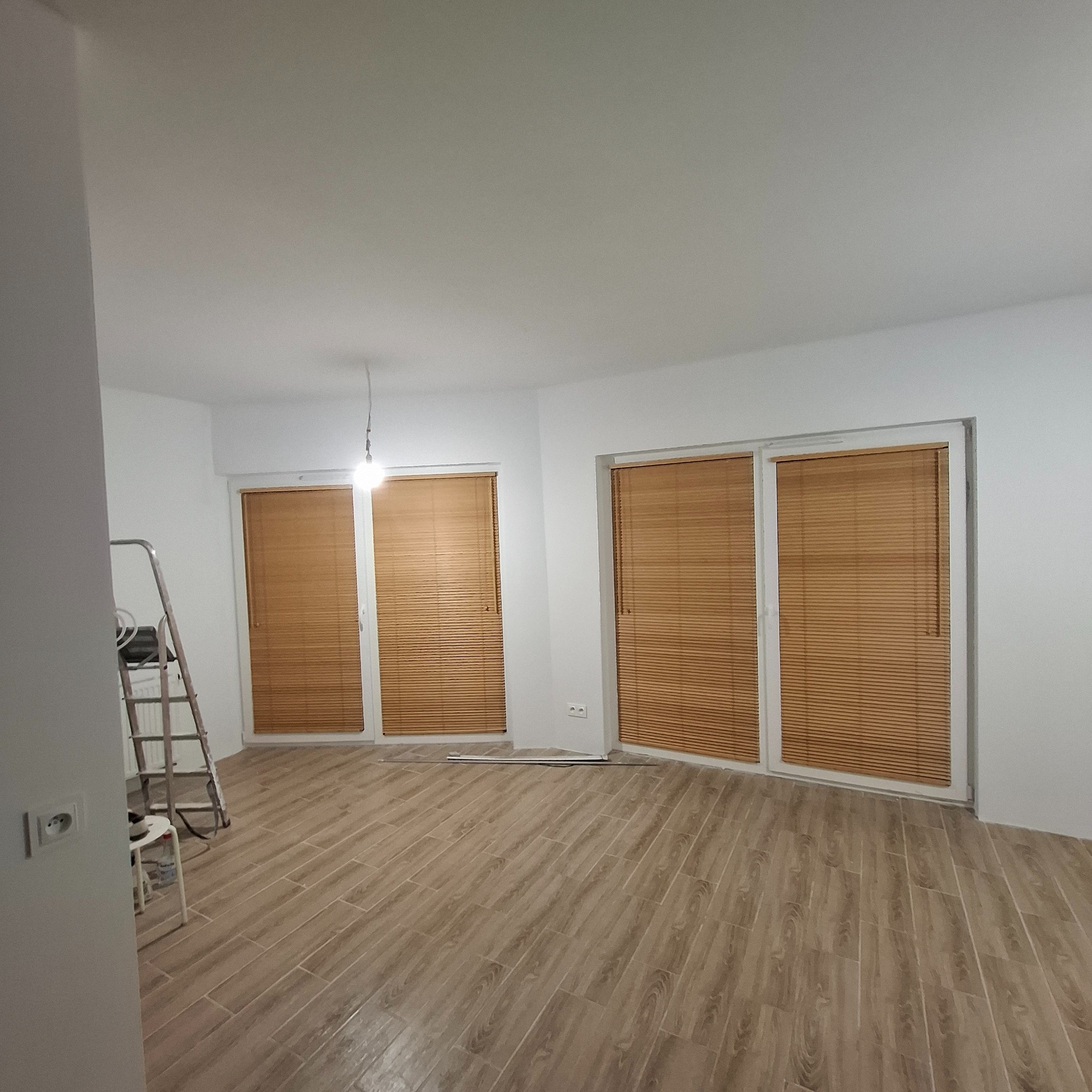 Jasne wnętrze po remoncie: świeżo pomalowane ściany, ułożona podłoga imitująca drewno, okna z drewnianymi żaluzjami. Widoczna drabina malarska.