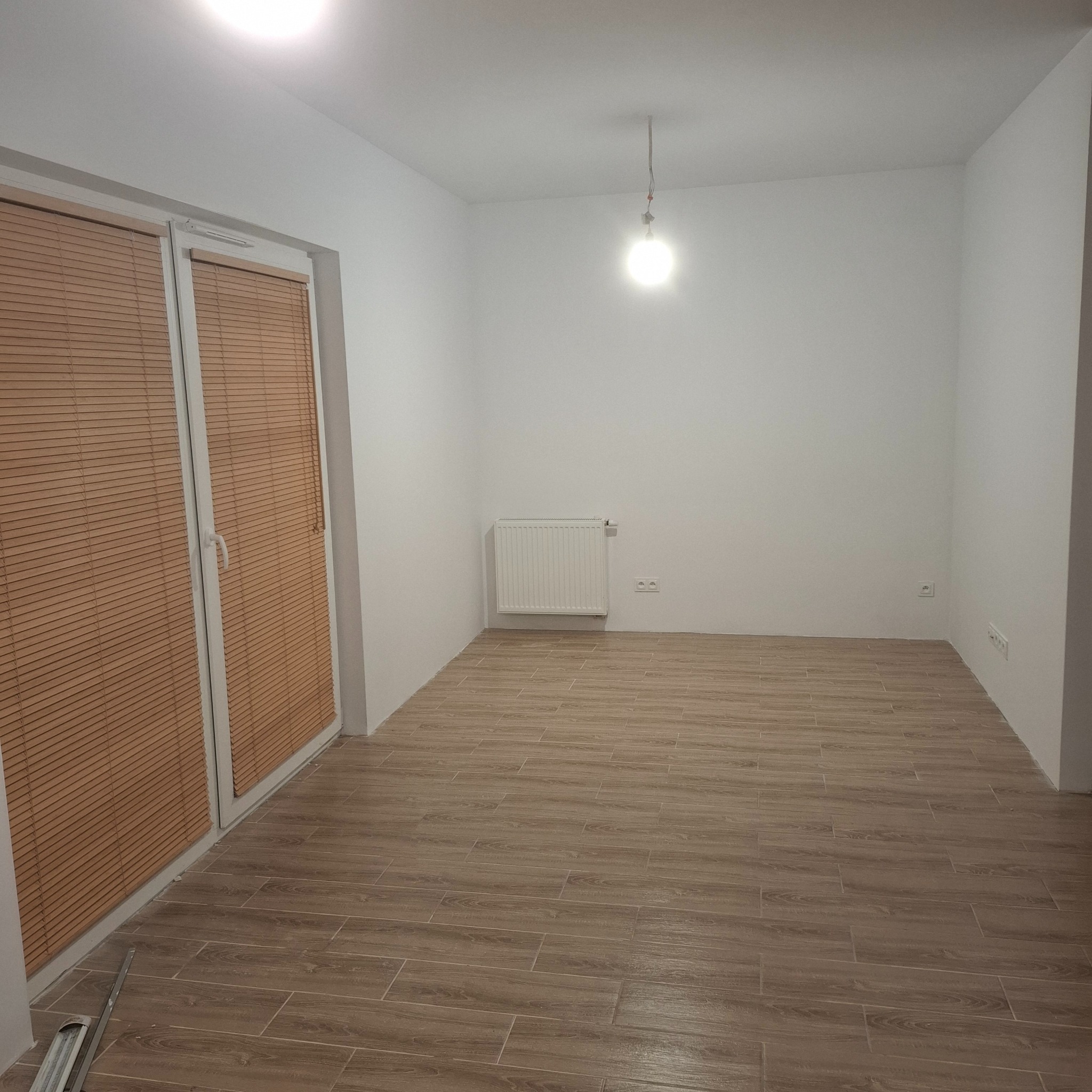 Jasne płytki podłogowe imitujące drewno w pustym pokoju z białymi ścianami, grzejnikiem i oknem z żaluzjami. Widoczna wisząca żarówka.